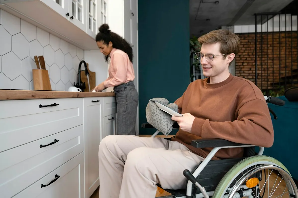Joven en silla de ruedas sonriendo en la cocina de su casa mientras una amiga prepara café